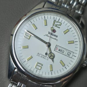 May include: Silver-toned Titoni Airmaster watch with a white face. The watch features a date display, luminous hands and markers, and a stainless steel band. The watch face displays the text "AUTOMATIC 25 JEWELS".