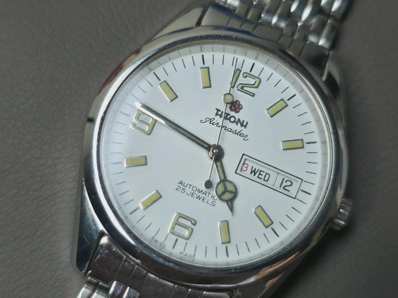 May include: Silver-toned Titoni Airmaster watch with a white face. The watch features a date display, luminous hands and markers, and a stainless steel band. The watch face displays the text "AUTOMATIC 25 JEWELS".