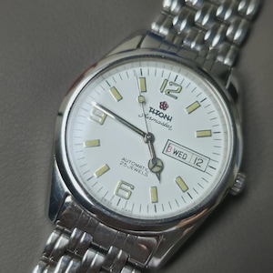 May include: Silver-toned wristwatch with a white face, featuring the brand name "Titoni Airmaster." The watch has a date display and luminous markers. The watch band is made of linked metal. The watch face displays the day and date.