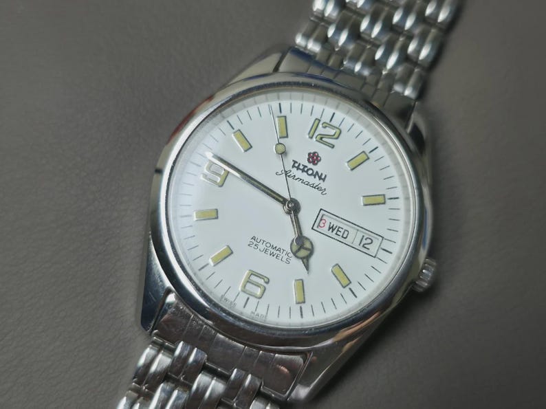 May include: Silver-toned wristwatch with a white face, featuring the brand name "Titoni Airmaster." The watch has a date display and luminous markers. The watch band is made of linked metal. The watch face displays the day and date.