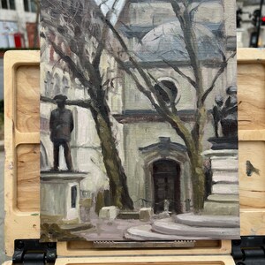 May include: An oil painting on canvas depicts a city scene with statues, trees, and a building. The artwork is displayed on a wooden easel. The color palette is muted, with shades of gray, brown, and beige. The painting captures a moment in an urban environment.