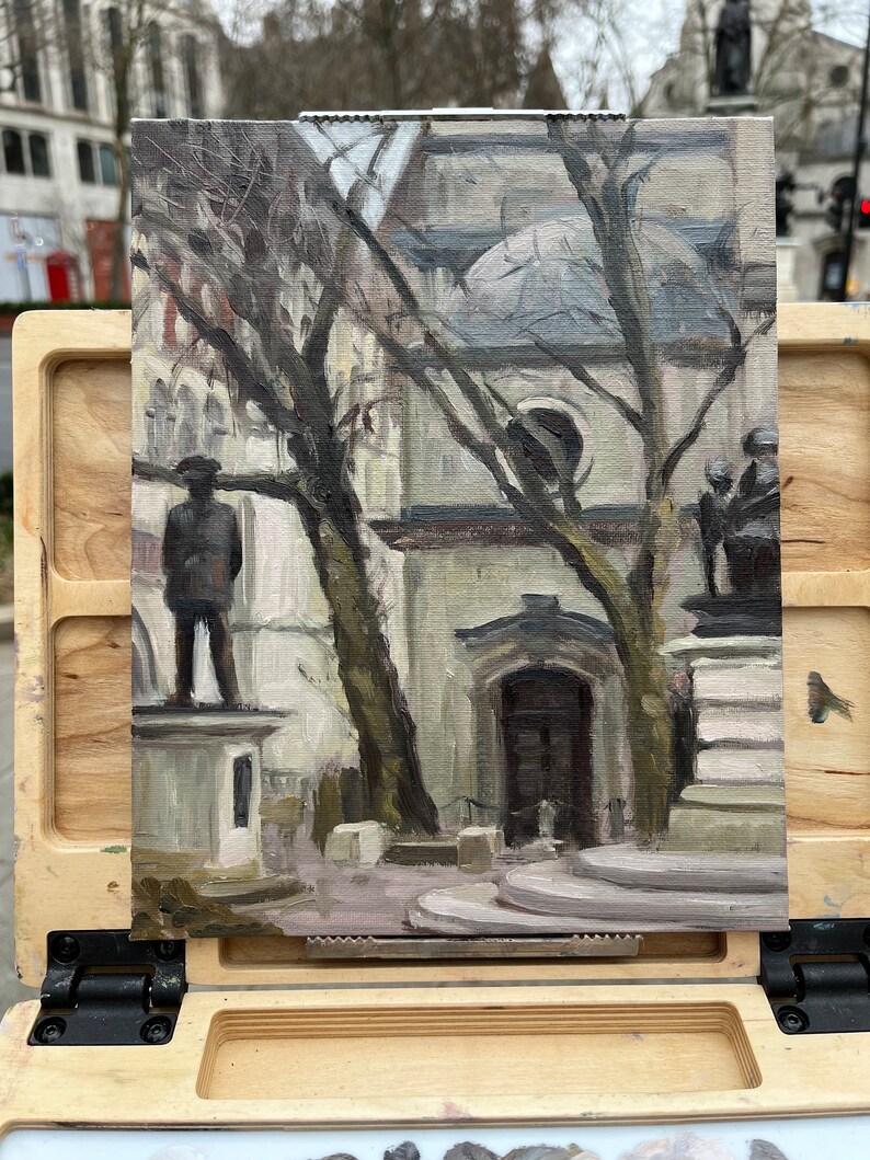 May include: An oil painting on canvas depicts a city scene with statues, trees, and a building. The artwork is displayed on a wooden easel. The color palette is muted, with shades of gray, brown, and beige. The painting captures a moment in an urban environment.