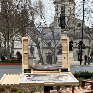 May include: An open wooden artist's easel with a painting of a building and statues in a public square. The easel has a built-in palette with mixed paint colors. The scene is outdoors on a cloudy day.