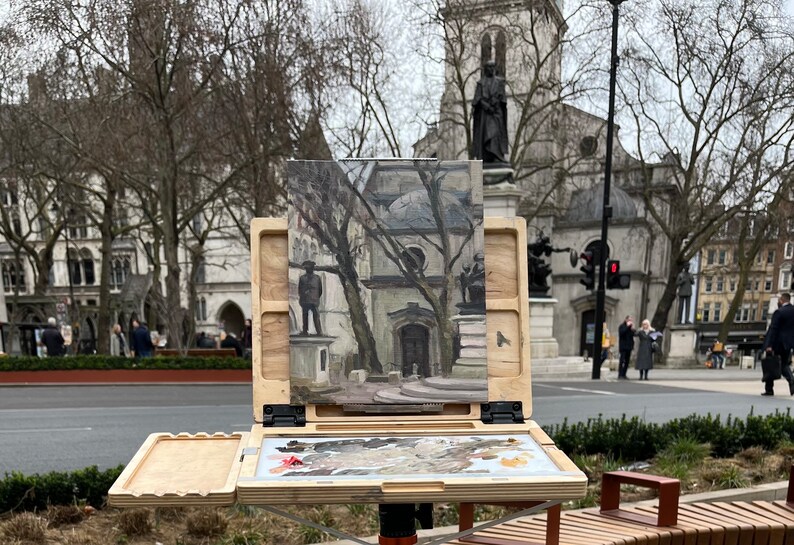 May include: An open wooden artist's easel with a painting of a building and statues in a public square. The easel has a built-in palette with mixed paint colors. The scene is outdoors on a cloudy day.