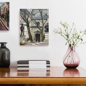 May include: A still life arrangement featuring two framed paintings, a pink glass vase with white flowers, and two decorative glass bottles. A black and white striped box sits on a wooden surface.