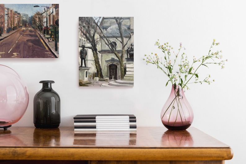 May include: A still life arrangement featuring two framed paintings, a pink glass vase with white flowers, and two decorative glass bottles. A black and white striped box sits on a wooden surface.