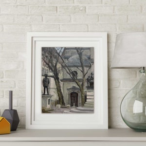 May include: A framed painting of a European city square with statues and bare trees. The artwork is in a white frame, displayed on a white shelf with decorative objects and a glass lamp with a white shade. The background is a white brick wall.