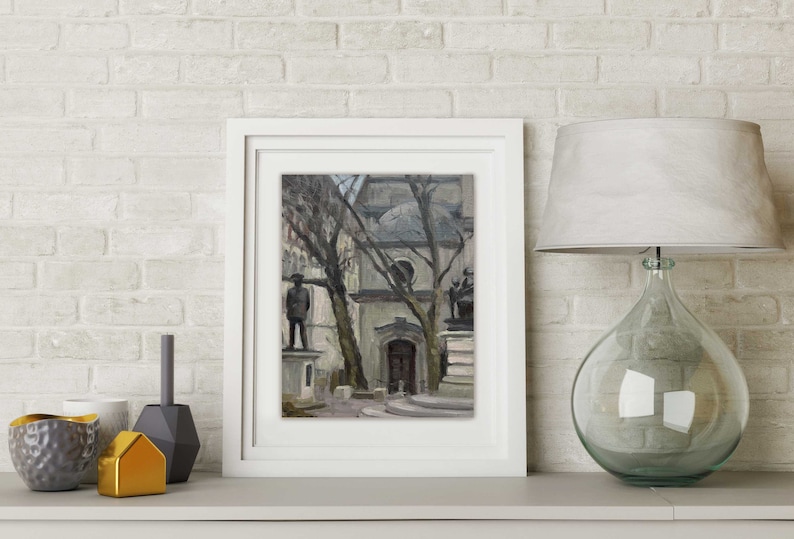 May include: A framed painting of a European city square with statues and bare trees. The artwork is in a white frame, displayed on a white shelf with decorative objects and a glass lamp with a white shade. The background is a white brick wall.