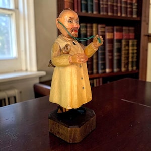 May include: A hand-carved wooden figurine of a doctor wearing a light yellow coat, standing on a dark brown octagonal base. The figure has a stethoscope around the neck, with one earpiece in the ear and the other held in the hand, featuring black shoes.