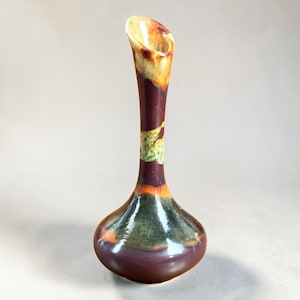 May include: A tall, slender vase with a flared opening. The vase has a dark burgundy base that transitions to a lighter brown and yellow at the top. A band of green and yellow accents the neck. The vase has a glossy finish.