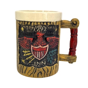 Könnte beinhalten: Ein Keramik-Bierkrug mit Holzoptik. Der Krug zeigt einen rot-weiß-blauen Adler mit einem Schild und dem Text "1792 INN". Der Henkel ist aus Holz mit einem roten Akzent.