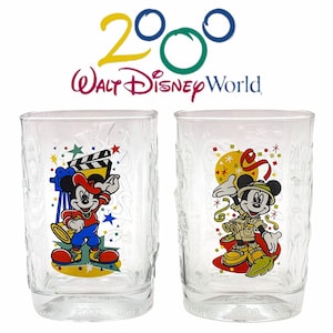 May include: Two clear glass tumblers with Mickey Mouse designs. The tumblers feature Mickey Mouse dressed as a filmmaker and Mickey Mouse dressed as an explorer. Both tumblers have the text "2000 Walt Disney World" on the bottom.