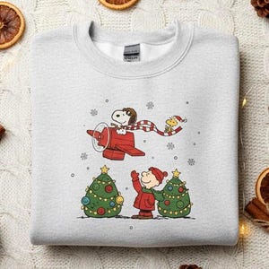 Peut inclure: Sweat-shirt gris clair avec un motif Peanuts sur le thème de Noël. Snoopy pilote un avion rouge avec Woodstock, et Charlie Brown est près d'un sapin de Noël. Décoré de pommes de pin, d'oranges séchées et de bâtons de cannelle.