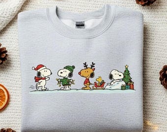May include: Ash-colored sweatshirt featuring a festive Snoopy and friends design. The graphic shows Snoopy, Woodstock, and other characters in holiday attire, with a Christmas tree and gifts. The text "color: ash" is in the upper left corner.