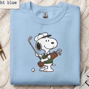 Peut inclure: Sweat-shirt bleu clair avec un personnage de dessin animé Snoopy jouant au golf, tenant un club et un sac de clubs. Le personnage porte un chapeau blanc et un gilet vert. Une balle de golf est au sol. Le texte "color: light blue" est en haut à gauche.