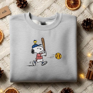 May include: Ash grey sweatshirt with a cartoon Snoopy character playing baseball. Snoopy, wearing a baseball cap and uniform, swings a bat at a yellow baseball. The sweatshirt has a crew neck design.