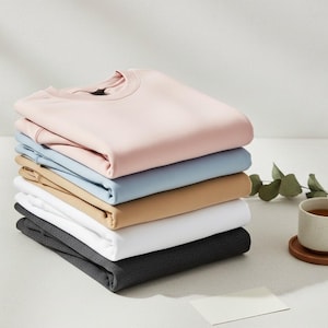 May include: A stack of six folded crew neck sweatshirts in various colors: light pink, light blue, tan, white, and dark gray. A cup of tea and a small card are on the table.
