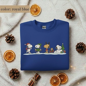 May include: Royal blue sweatshirt featuring a cartoon design of Snoopy and friends in winter attire. The graphic includes Snoopy wearing a Santa hat, a green-clad Woodstock, and other characters. The sweatshirt is a crew neck style.