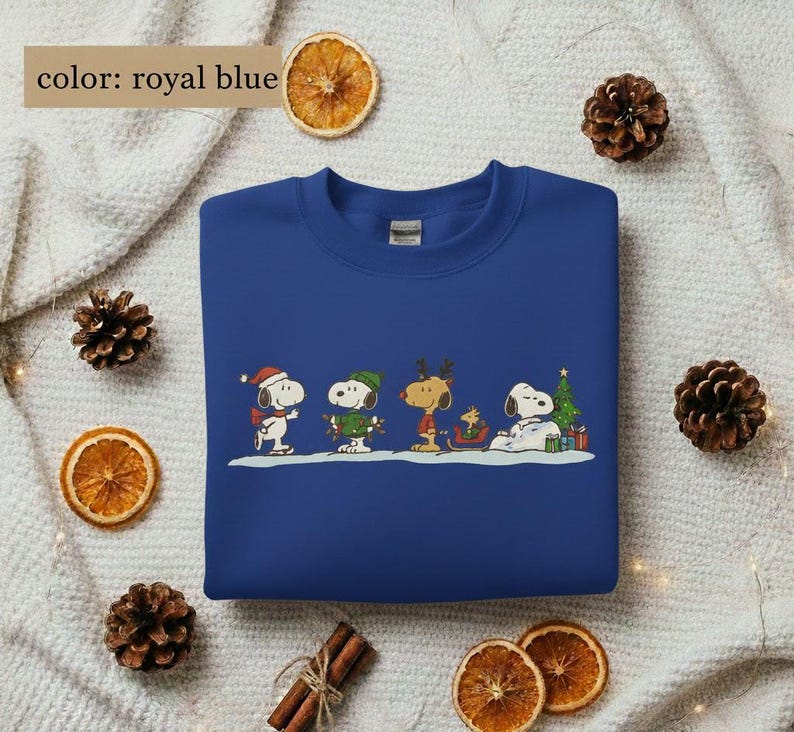 May include: Royal blue sweatshirt featuring a cartoon design of Snoopy and friends in winter attire. The graphic includes Snoopy wearing a Santa hat, a green-clad Woodstock, and other characters. The sweatshirt is a crew neck style.