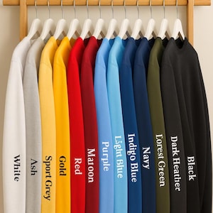 May include: A display of crewneck sweatshirts in various colors, including white, ash, sport grey, gold, red, maroon, purple, light blue, indigo blue, navy, forest green, dark heather, and black. Each sweatshirt hangs on a wooden rack.