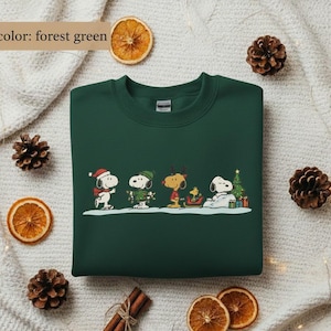 May include: Forest green sweatshirt featuring a festive Snoopy and Peanuts gang design. The graphic shows Snoopy and friends in winter attire, including a Santa hat, scarf, and reindeer antlers. A Christmas tree and presents are also included in the design.