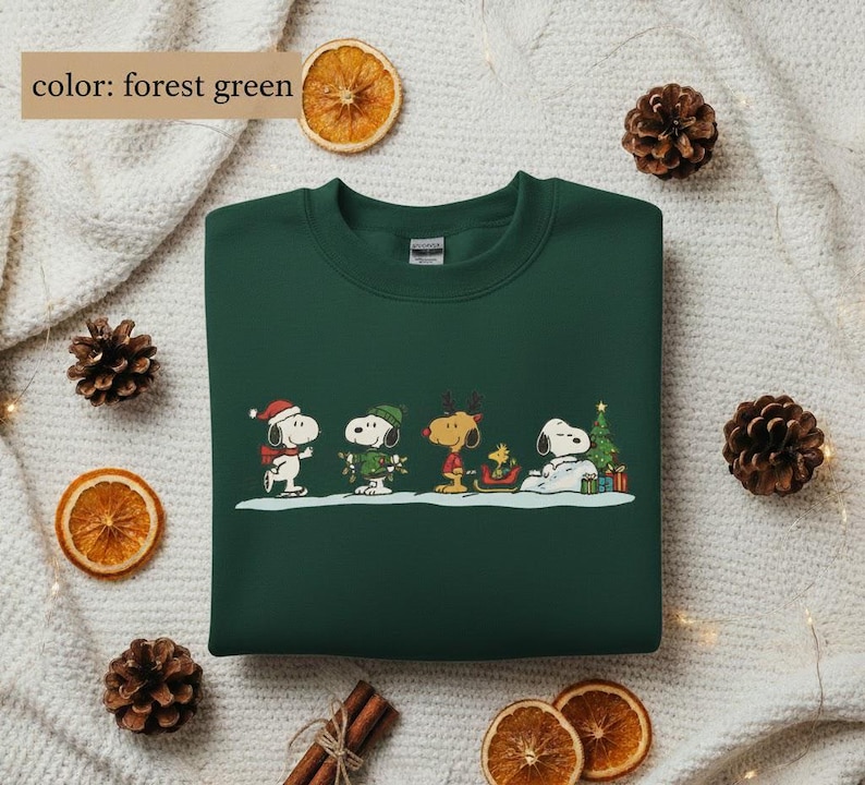 May include: Forest green sweatshirt featuring a festive Snoopy and Peanuts gang design. The graphic shows Snoopy and friends in winter attire, including a Santa hat, scarf, and reindeer antlers. A Christmas tree and presents are also included in the design.