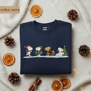May include: Navy blue sweatshirt featuring a festive Snoopy and friends design. The graphic shows Snoopy, Woodstock, and other Peanuts characters in winter attire, with a Christmas tree and gifts. The sweatshirt is surrounded by pine cones and dried orange slices.