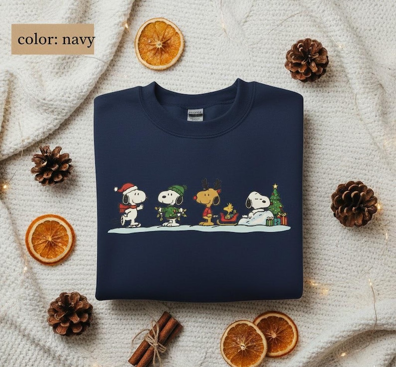 May include: Navy blue sweatshirt featuring a festive Snoopy and friends design. The graphic shows Snoopy, Woodstock, and other Peanuts characters in winter attire, with a Christmas tree and gifts. The sweatshirt is surrounded by pine cones and dried orange slices.