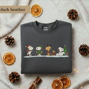 May include: Dark heather gray sweatshirt featuring a colorful graphic of Snoopy and friends in holiday attire. The design includes Snoopy wearing a Santa hat, a green-clad Woodstock, and other Peanuts characters. The text "dark heather" is in the upper left corner.