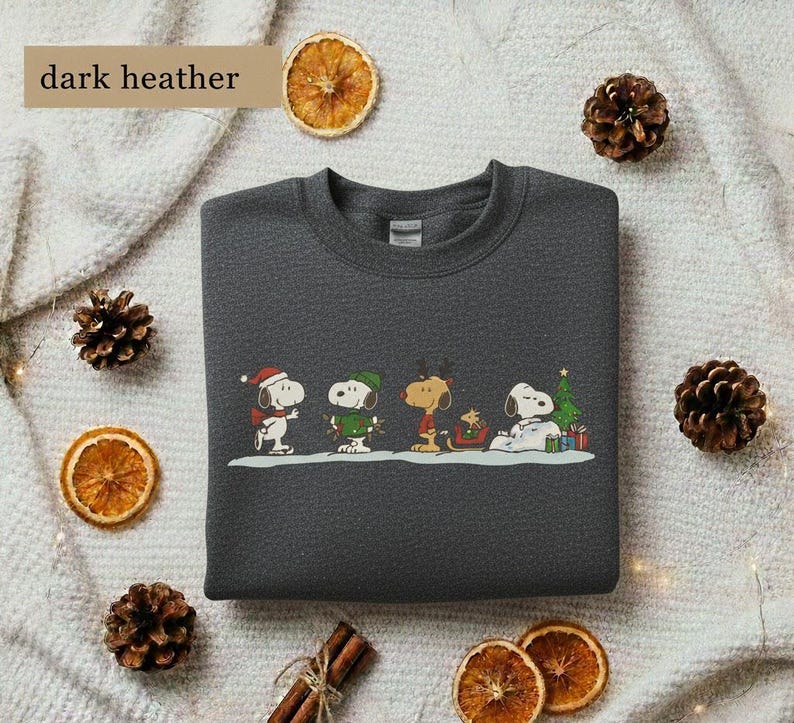 May include: Dark heather gray sweatshirt featuring a colorful graphic of Snoopy and friends in holiday attire. The design includes Snoopy wearing a Santa hat, a green-clad Woodstock, and other Peanuts characters. The text "dark heather" is in the upper left corner.