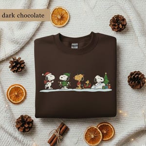 May include: Dark chocolate-colored sweatshirt featuring a festive Snoopy and friends design. The graphic includes Snoopy in a Santa hat, Woodstock, and other characters in winter attire. The text "dark chocolate" is in the upper left corner.