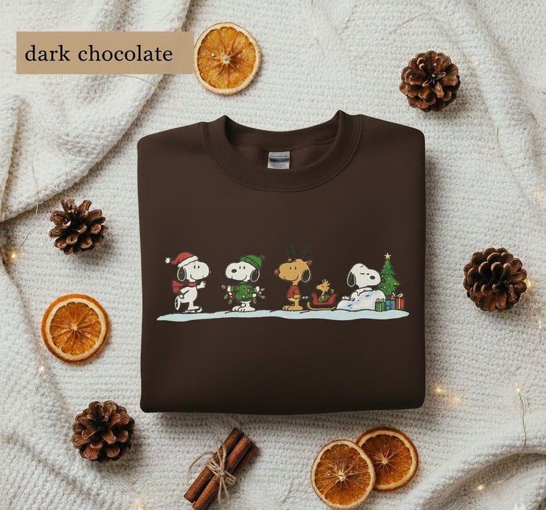 May include: Dark chocolate-colored sweatshirt featuring a festive Snoopy and friends design. The graphic includes Snoopy in a Santa hat, Woodstock, and other characters in winter attire. The text "dark chocolate" is in the upper left corner.