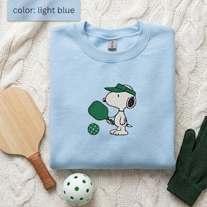 Peut inclure: Sweat-shirt bleu clair avec un personnage de dessin animé jouant au pickleball. Le motif comprend une raquette, une balle et une visière vertes. Une raquette en bois, un gant vert et une balle blanche à pois verts sont également visibles.