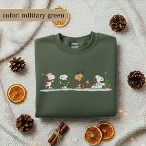 May include: Military green sweatshirt featuring a festive Snoopy and friends design. The graphic includes Snoopy in a Santa hat, a green elf hat, reindeer antlers, and a Christmas tree. The sweatshirt is a crew neck style.