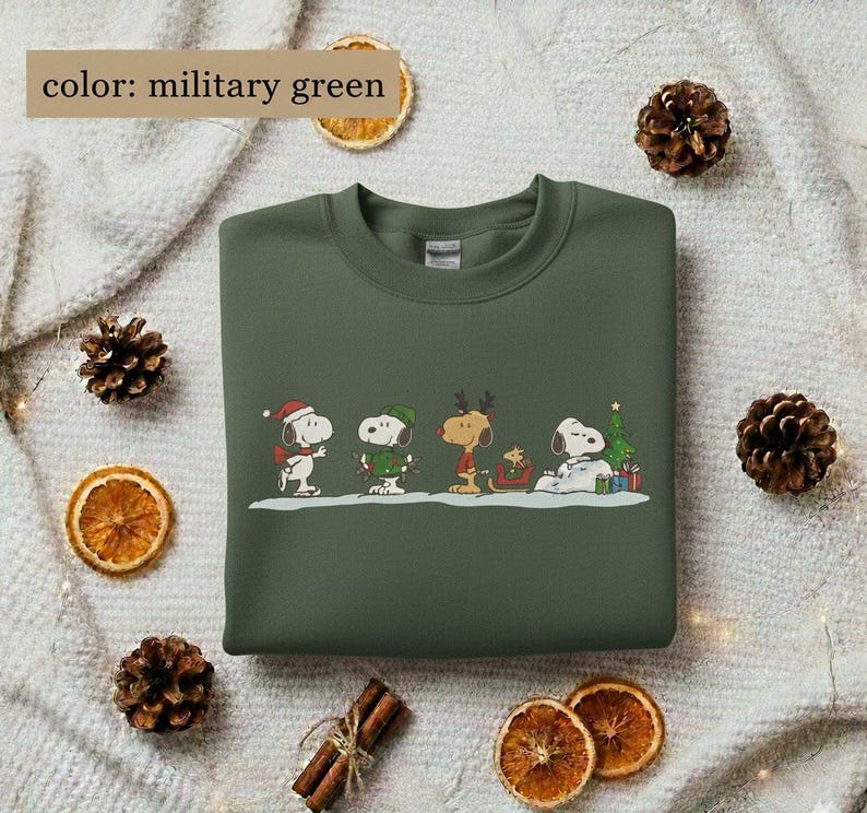 May include: Military green sweatshirt featuring a festive Snoopy and friends design. The graphic includes Snoopy in a Santa hat, a green elf hat, reindeer antlers, and a Christmas tree. The sweatshirt is a crew neck style.