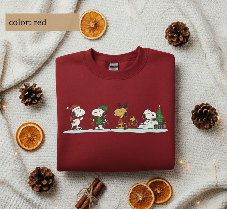 May include: A red crewneck sweatshirt featuring a festive Snoopy and friends design. The graphic includes Snoopy, Woodstock, and other Peanuts characters in holiday attire, with a Christmas tree and gifts. The text "color: red" is in the upper left corner.