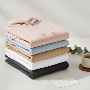 May include: A stack of six folded hoodies in various colors: light pink, light blue, tan, white, and dark gray. The top hoodie is light pink with a hood and drawstring. A cup of tea and a blank card are on the side.