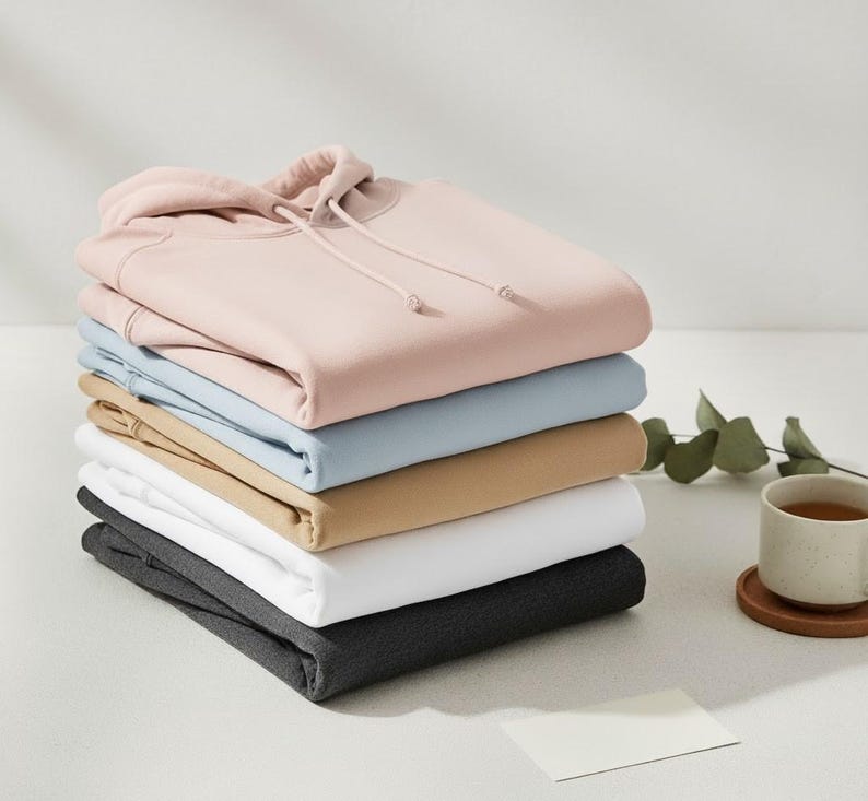 May include: A stack of six folded hoodies in various colors: light pink, light blue, tan, white, and dark gray. The top hoodie is light pink with a hood and drawstring. A cup of tea and a blank card are on the side.