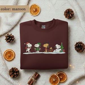 May include: Maroon sweatshirt featuring a festive Snoopy and friends design. The graphic includes Snoopy in a Santa hat, Woodstock, and other characters in winter attire. The text "color: maroon" is in the upper left corner. The sweatshirt is surrounded by pinecones and dried orange slices.