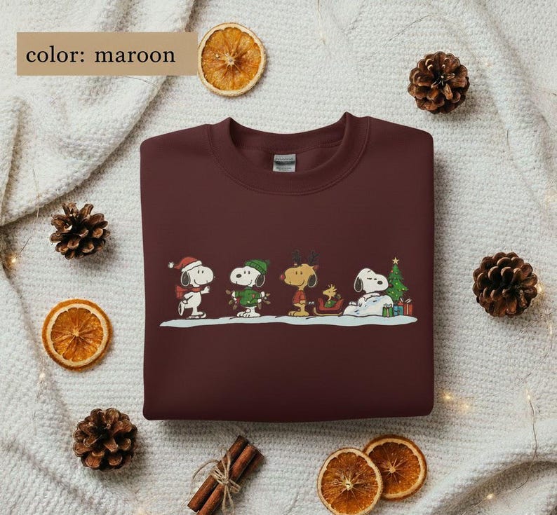 May include: Maroon sweatshirt featuring a festive Snoopy and friends design. The graphic includes Snoopy in a Santa hat, Woodstock, and other characters in winter attire. The text "color: maroon" is in the upper left corner. The sweatshirt is surrounded by pinecones and dried orange slices.