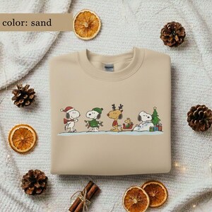 May include: Sand-colored sweatshirt featuring a festive Snoopy and friends design. The graphic includes Snoopy in a Santa hat, a green-clad Snoopy, a reindeer, and Snoopy resting by a Christmas tree. The sweatshirt is surrounded by decorative elements.