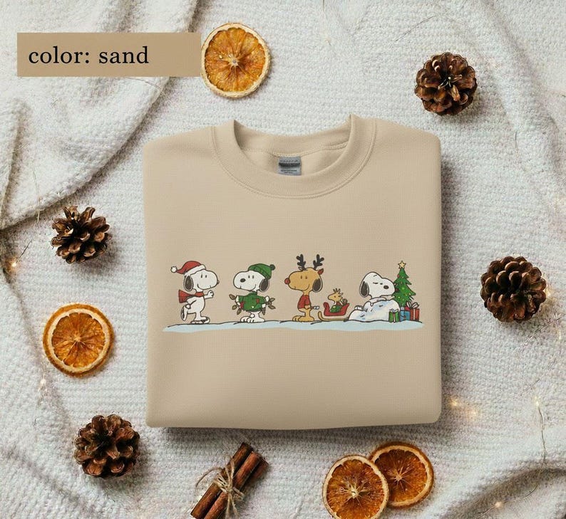 May include: Sand-colored sweatshirt featuring a festive Snoopy and friends design. The graphic includes Snoopy in a Santa hat, a green-clad Snoopy, a reindeer, and Snoopy resting by a Christmas tree. The sweatshirt is surrounded by decorative elements.