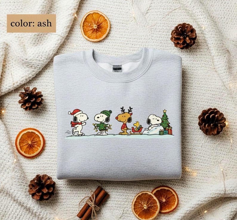 May include: Ash-colored sweatshirt featuring a festive Snoopy and friends design. The graphic shows Snoopy, Woodstock, and other characters in holiday attire, with a Christmas tree and gifts. The text "color: ash" is in the upper left corner.