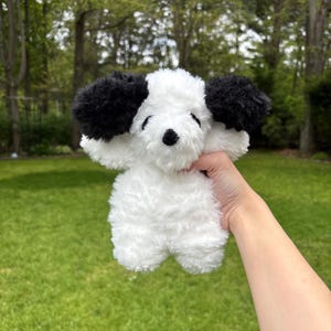 May include: A plush white dog toy with black ears and a black nose is held up against a green background. The toy is soft and fluffy, with a friendly expression. The toy is designed to look like a puppy.