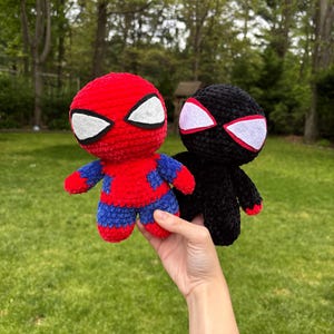May include: Two crocheted superhero dolls. One is red and blue with white eye shapes, the other is black with white and red eye shapes. The dolls are held in front of a green background.