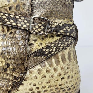 Puede incluir: Primer plano de una bota de piel de serpiente con suela de cuero marr&oacute;n. La bota presenta una correa con una hebilla plateada. El estampado de piel de serpiente incluye tonos de marr&oacute;n, beige y crema.