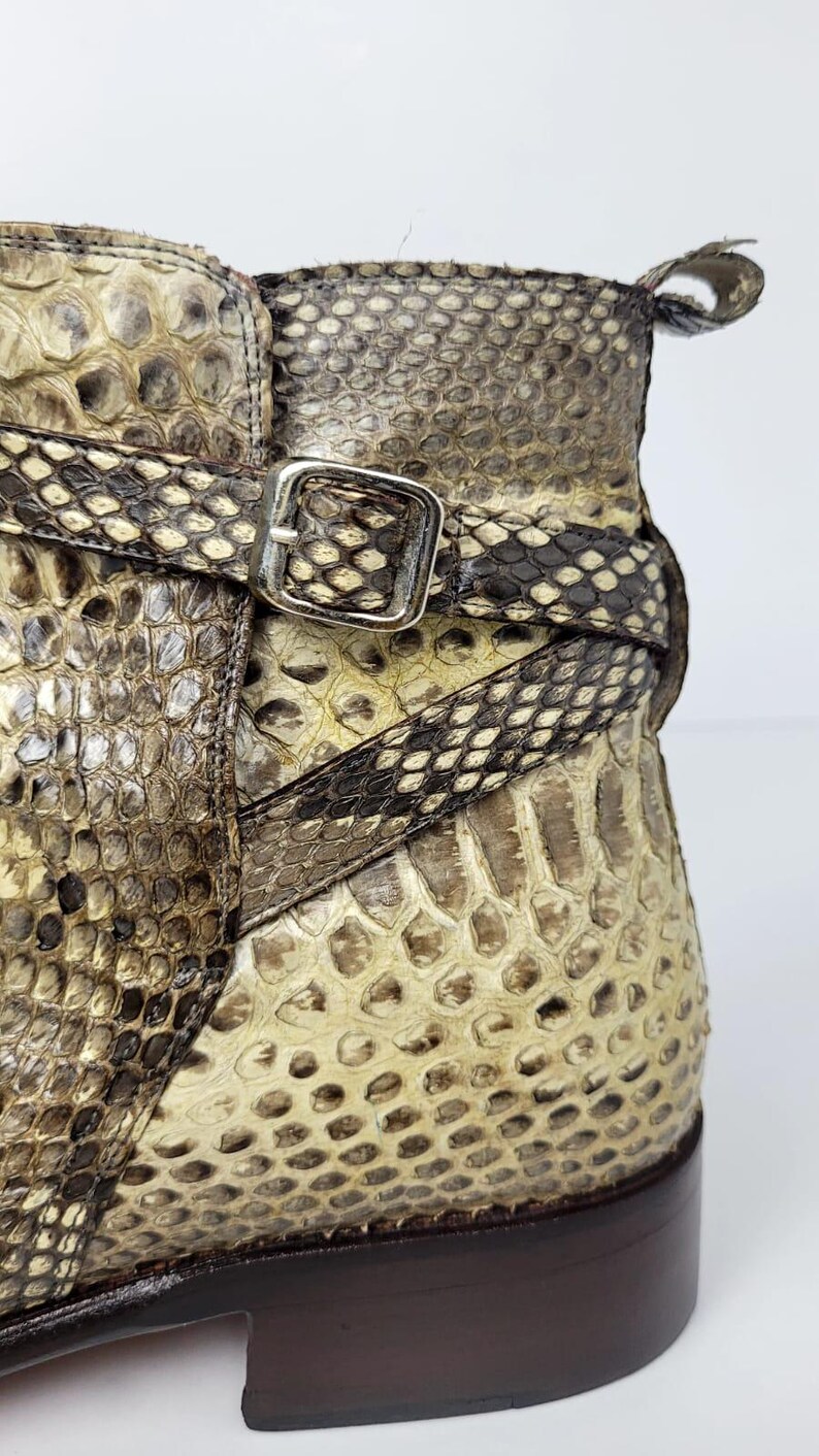 Puede incluir: Primer plano de una bota de piel de serpiente con suela de cuero marr&oacute;n. La bota presenta una correa con una hebilla plateada. El estampado de piel de serpiente incluye tonos de marr&oacute;n, beige y crema.
