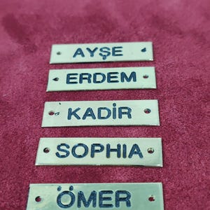 Puede incluir: Cinco etiquetas rectangulares de lat&oacute;n con letras negras. Se muestran los nombres Ayse, Erdem, Kadir, Sophia y Omer. Cada etiqueta tiene dos peque&ntilde;os agujeros y est&aacute; sobre un fondo burdeos.