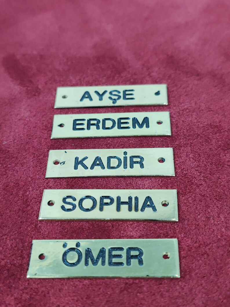 Puede incluir: Cinco etiquetas rectangulares de lat&oacute;n con letras negras. Se muestran los nombres Ayse, Erdem, Kadir, Sophia y Omer. Cada etiqueta tiene dos peque&ntilde;os agujeros y est&aacute; sobre un fondo burdeos.