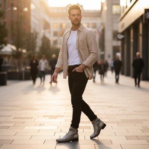 Puede incluir: Un hombre con una camisa beige sobre una camiseta blanca, pantalones negros y botines estampados camina por una calle de la ciudad. El fondo muestra peatones y edificios borrosos, con luz solar.